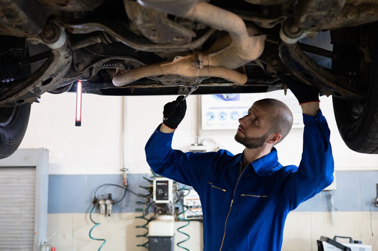 About A mechanic in blue overalls inspects the exhaust system of a car in a workshop.